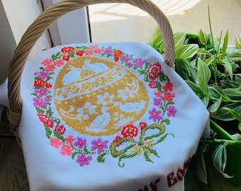 May include: White fabric with a cross-stitch design of a golden Easter egg surrounded by colourful flowers. The text "Веселих Воскресень" is embroidered in brown at the bottom of the fabric.