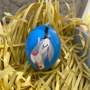 Puede incluir: Un huevo de Pascua azul con una imagen pintada de un conejo gris con un sombrero de copa y una corbata de lazo. El huevo está sentado en un nido de papel amarillo triturado.