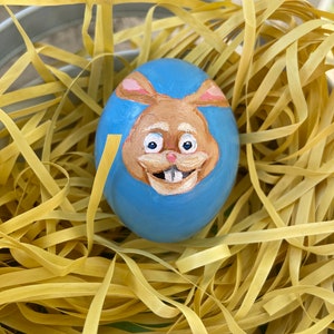 Puede incluir: Un huevo de Pascua azul con una cara de conejo pintada. El conejo tiene pelaje marrón, orejas rosas y una sonrisa blanca. El huevo está en un nido de paja amarilla.