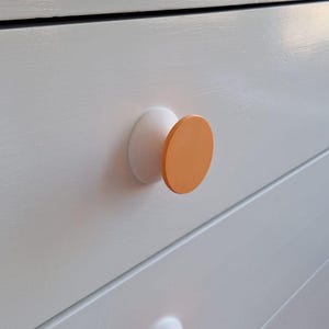 Peut inclure: Une commode blanche dotée de plusieurs tiroirs horizontaux aux surfaces lisses. Chaque tiroir est équipé d'un bouton rond, dont la base est blanche et la face avant est un disque circulaire orange vif et uni, créant un contraste marqué.