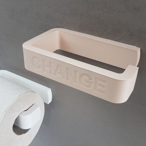 Può includere: Un portarotolo di carta igienica beige chiaro con la scritta "CHANGE" in rilievo sul davanti. Un portarotolo di carta igienica bianco è attaccato a sinistra, con un rotolo di carta igienica bianca. Gli oggetti sono montati su un muro grigio.