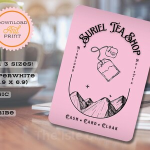 May include: A pink printable card with a black and white illustration of a tea bag, mountains, and stars. The text "SURIEL TEA SHOP" is in a black cursive font. The text "HIGHLORD HOT TEA" is in a smaller black font. The text "CASH + CARD + CLOAK" is in a smaller black font.