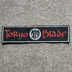 Puede incluir: Un parche rectangular negro con las palabras "Tokyo Blade" en rojo, con letras estilizadas. Un emblema circular con un diseño blanco y negro está centrado entre las palabras. El parche es probablemente para ropa o accesorios.