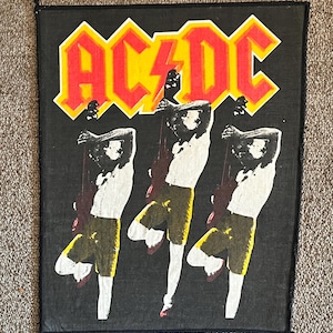 May include: Black rectangular patch featuring the band name "AC/DC" in red letters with a yellow outline. Below the text are three stylized figures playing guitars. The figures are in white, yellow, and black.