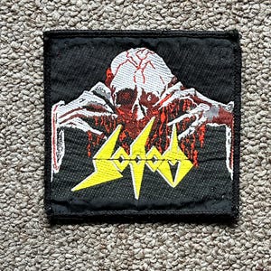 Könnte beinhalten: Schwarzer quadratischer Aufnäher mit einem Schädel in Rot und Weiß und dem gelben Wort "SODOM". Der Aufnäher hat einen schwarzen Rand und ist wahrscheinlich für Kleidung oder Accessoires.