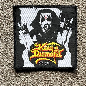 May include: Square black and white patch featuring a graphic of King Diamond, with the band's logo in yellow and red, and the name "Abigail" below. The patch has a black border.