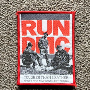 Puede incluir: Parche bordado con borde rojo. Presenta las palabras "RUN DMC" en letras grandes de color naranja, una imagen en blanco y negro del grupo y el texto "TOUGHER THAN LEATHER".
