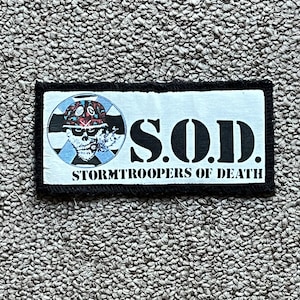 May include: A rectangular patch with the text "S.O.D. STORMTROOPERS OF DEATH" in black block letters. The patch features a skull wearing a helmet with a red and blue design. The patch has a black border.