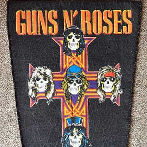 May include: A black fabric patch with the words "Guns N' Roses" in orange lettering. Five skull faces are arranged in a cross pattern, with each skull wearing a different hat.