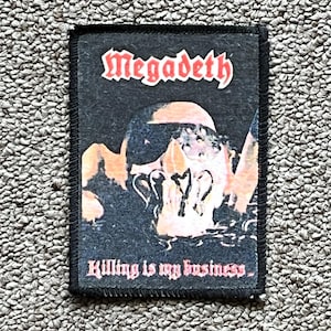 May include: Black rectangular patch with the band name "Megadeth" in red. The patch features a skull graphic and the text "Killing is my business" in pink. The patch has a black border and is designed for clothing.