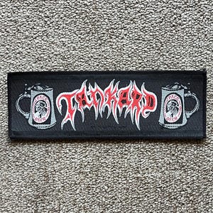 May include: Black rectangular patch featuring the word "TANKARD" in red, spiky lettering. Two beer mugs with "SPACE BEER" text and a skull graphic are on either side. The patch is on a textured, beige surface.