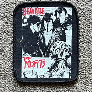 Może przedstawiać: Czarna prostokątna naszywka z obszytą krawędzią. Naszywka przedstawia szary obraz zespołu z czerwonym tekstem "BEWARE" na górze i "THE MISFITS" na dole. Obraz zawiera czaszkę.