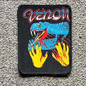 Puede incluir: Parche negro con la palabra "VENOM" en letras rojas y blancas. Una serpiente azul con detalles rojos y manos amarillas está en el centro. El parche tiene un borde negro.