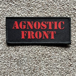 Könnte beinhalten: Ein schwarzer rechteckiger Aufnäher mit den Worten "AGNOSTIC FRONT" in Rot gestickt. Der Aufnäher ist wahrscheinlich aus Stoff und hat einen genähten Rand. Der Hintergrund ist eine strukturierte, neutralfarbene Oberfläche.