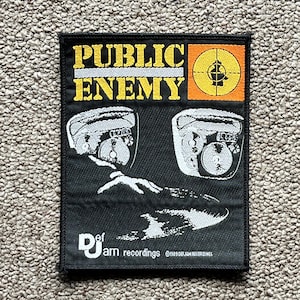 May include: Black Public Enemy patch featuring the band's name in yellow, a target logo, and two boombox illustrations. The patch also includes "Def Jam recordings" and copyright information.