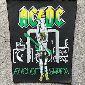 Pode incluir: Patch preto com o logotipo AC/DC em amarelo e verde, e o texto "FLICK OF THE SWITCH" em verde. O patch mostra um gráfico de palco e uma figura segurando uma guitarra verde.