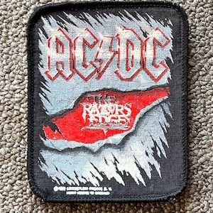 May include: Black and white woven patch with the band name "AC/DC" in red lettering. The patch features a red and white graphic of a hand reaching out from a rock formation with the words "The Razor's Edge" in white lettering.
