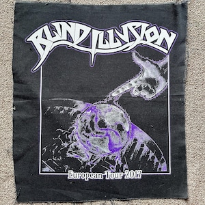 May include: A black fabric patch featuring a purple and white graphic of a hand reaching toward a globe. The text BLIND ILLUSION is printed in a stylized font at the top, and the text European Tour 2017 is printed in white at the bottom.