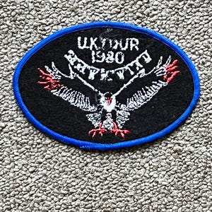 May include: Black oval patch with a blue border featuring a white eagle with outstretched wings and red accents. The text "UK TOUR 1980" is arched above the eagle, indicating a tour souvenir or memorabilia item.