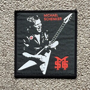 Puede incluir: Parche cuadrado negro con una ilustración blanca de un guitarrista tocando una guitarra en forma de V. El parche tiene el nombre "MICHAEL SCHENKER" en texto rojo en la parte superior y un logotipo rojo en la parte inferior.
