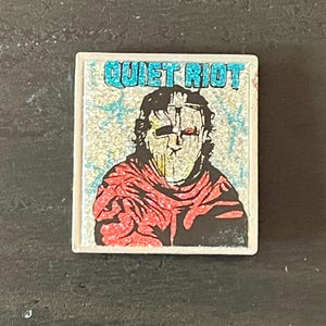 May include: A white and blue sticker with a black and white illustration of a person wearing a red and black mask. The sticker says "Quiet Riot" in blue letters.
