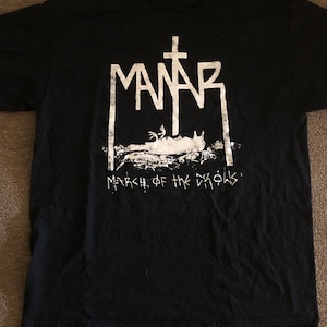 May include: Black t-shirt with a white graphic featuring the word "MANAR" above a cross and a stylized illustration. The text "MARCH OF THE CROWS" is below the illustration. The shirt is made of a soft material.