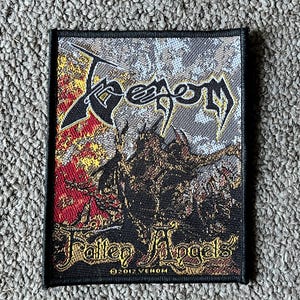 May include: A rectangular woven fabric patch featuring the Venom logo at the top and the text Fallen Angels at the bottom. The design includes a dark central figure against a background of red, yellow, and grey textures with a black border.
