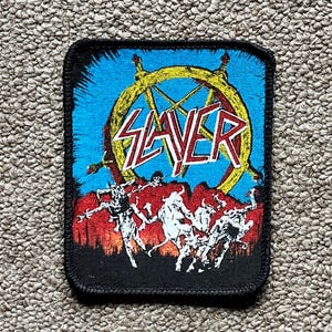 May include: A rectangular embroidered patch featuring the Slayer logo in white with red outlines, set against a yellow pentagram and a bright blue sky. Below, white skeletal figures are depicted against a dark red and black landscape background.