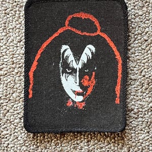 Pode incluir: Patch bordado preto e vermelho com um rosto estilizado com maquiagem branca e um halo vermelho.