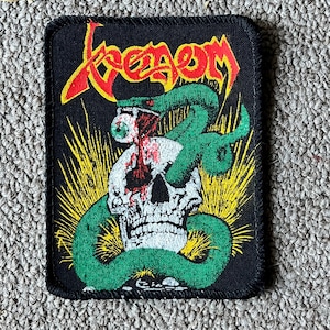 May include: A rectangular black fabric patch featuring the word Venom in red and yellow lettering. The design includes a white human skull with a green snake coiled around it, with the snake biting into a single eyeball dripping red blood.
