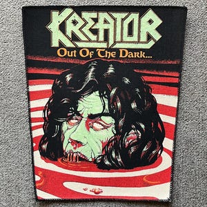 Op de afbeelding: Zwarte patch met de bandnaam "Kreator" in groen en de zin "Out Of The Dark..." in oranje. De patch toont een zombie-achtig gezicht dat opduikt uit rood-wit gestreept water.