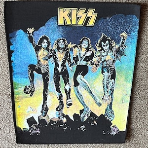 Pode incluir: Um remendo de tecido preto com uma imagem colorida da banda de rock KISS. Os membros da banda estão com sua maquiagem e figurinos característicos, e o nome da banda está impresso em letras amarelas na parte superior do remendo.