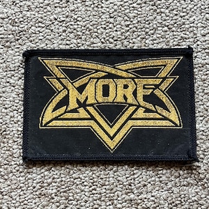Puede incluir: Parche rectangular negro con texto bordado y diseño geométrico en dorado. La palabra "MORE" está en dorado, rodeada por un patrón en forma de estrella. El parche es probablemente para ropa o accesorios.