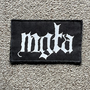 Pode incluir: Um patch retangular preto com a palavra "mota" em letras góticas brancas. O patch é feito de tecido tecido e provavelmente foi projetado para ser costurado ou passado a ferro em roupas ou acessórios.