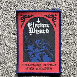 May include: A rectangular patch with a red border and a blue stitched edge. The patch features the words "Electric Wizard" in white gothic lettering, a blue illustration, and the text "Invoking Drugs and Murder" in red.
