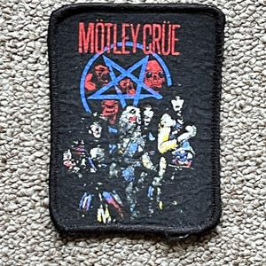May include: Black rectangular patch featuring the band name "Mötley Crüe" in red letters. Below the text is a blue pentagram with skulls and band members. The patch has a black border and is likely for a jacket or bag.
