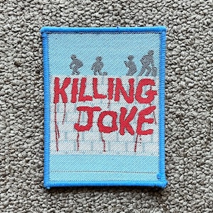 May include: A rectangular patch with a blue border and a light blue background. The words "KILLING JOKE" are in red, with a stylized font. Above the text are three silhouetted figures. The patch is on a textured, neutral-colored surface.