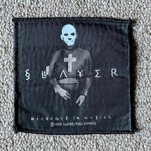 May include: A square black fabric patch featuring a white graphic of a figure with a cross on their chest. The text SLAYER is centered, with DIABOLUS IN MUSICA and 1998 SLAYER/ROLL EXPRESS printed below. The patch has a stitched border.