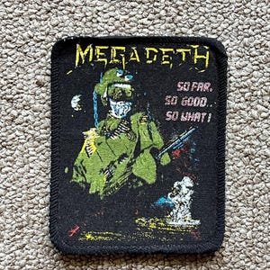 Puede incluir: Parche negro con el nombre de la banda "MEGADETH" en letras amarillas. El parche presenta una ilustración de una figura esquelética verde con uniforme militar, sosteniendo un arma. El texto en el parche dice "SO FAR, SO GOOD.. SO WHAT!"