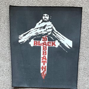 Puede incluir: Un parche cuadrado negro con una ilustración blanca de una figura sobre una cruz roja. Las palabras "BLACK SABBATH" están impresas en rojo en la cruz. El parche está diseñado para coserlo en ropa o bolsos.