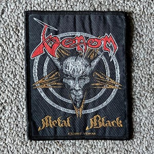 May include: A rectangular black embroidered patch featuring the red and white Venom logo at the top, a central grey goat head graphic within a circular design, and the gold text Metal Black at the bottom with the small text 2007 VENOM below it.