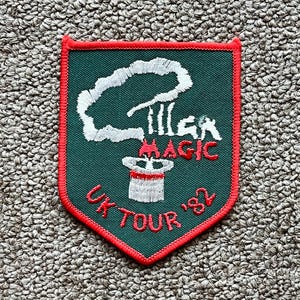 May include: Embroidered patch with a green background and a red border. The patch features a white smoke design, a top hat, and the words "Magic UK Tour '82" in red. The patch is likely a souvenir or collectible item.