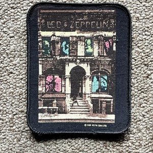 Puede incluir: Un parche rectangular negro con la portada del álbum Led Zeppelin. La imagen muestra un edificio con imágenes coloridas en las ventanas. El texto "LED ZEPPELIN" está en la parte superior. El parche tiene un borde negro y está sobre una superficie texturizada.