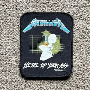 Puede incluir: Parche cuadrado negro con el nombre de la banda "Metallica" en letras azules y plateadas. El parche presenta un gráfico de una figura con una espada y el texto "Metal Up Your Ass".