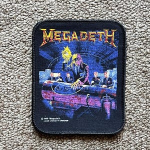 Pode incluir: Patch retangular preto com o nome da banda "MEGADETH" em letras douradas. O patch retrata uma cena com figuras em volta de uma mesa, com uma figura central segurando um objeto brilhante. O fundo é azul escuro.