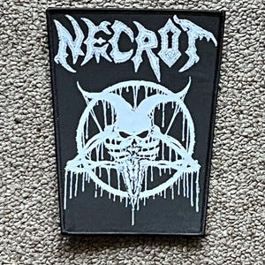 May include: Black rectangular patch with the word "NECROT" in white gothic lettering. Below the text is a white illustration of a skull within a pentagram, with dripping accents. The patch is likely for a jacket or bag.