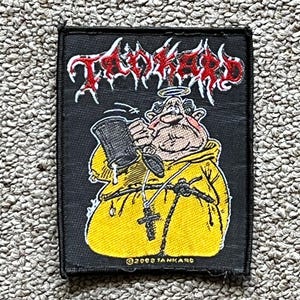 May include: Black rectangular patch featuring the word "Tankard" in red, gothic-style lettering. The patch depicts a cartoon monk in a yellow robe holding a beer stein. A halo is above the monk's head, and a cross hangs from his neck. The text "©2008 TANKARD" is at the bottom.