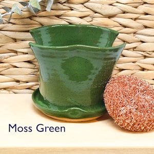 May include: A moss green ceramic planter with a wavy rim. The planter is sitting on a wooden surface next to a copper-colored scouring pad.