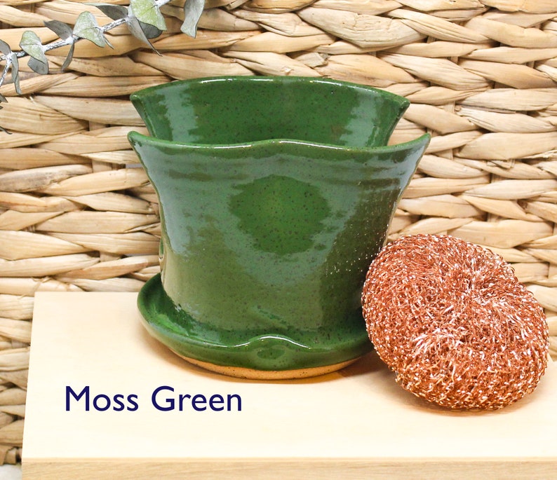 May include: A moss green ceramic planter with a wavy rim. The planter is sitting on a wooden surface next to a copper-colored scouring pad.