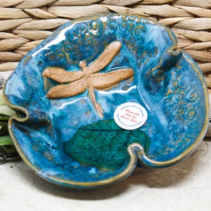 May include: A blue ceramic dish with a dragonfly design. The dish has a wavy edge and a green glaze in the center. The dish is handmade with lake-fired earthenware glass.